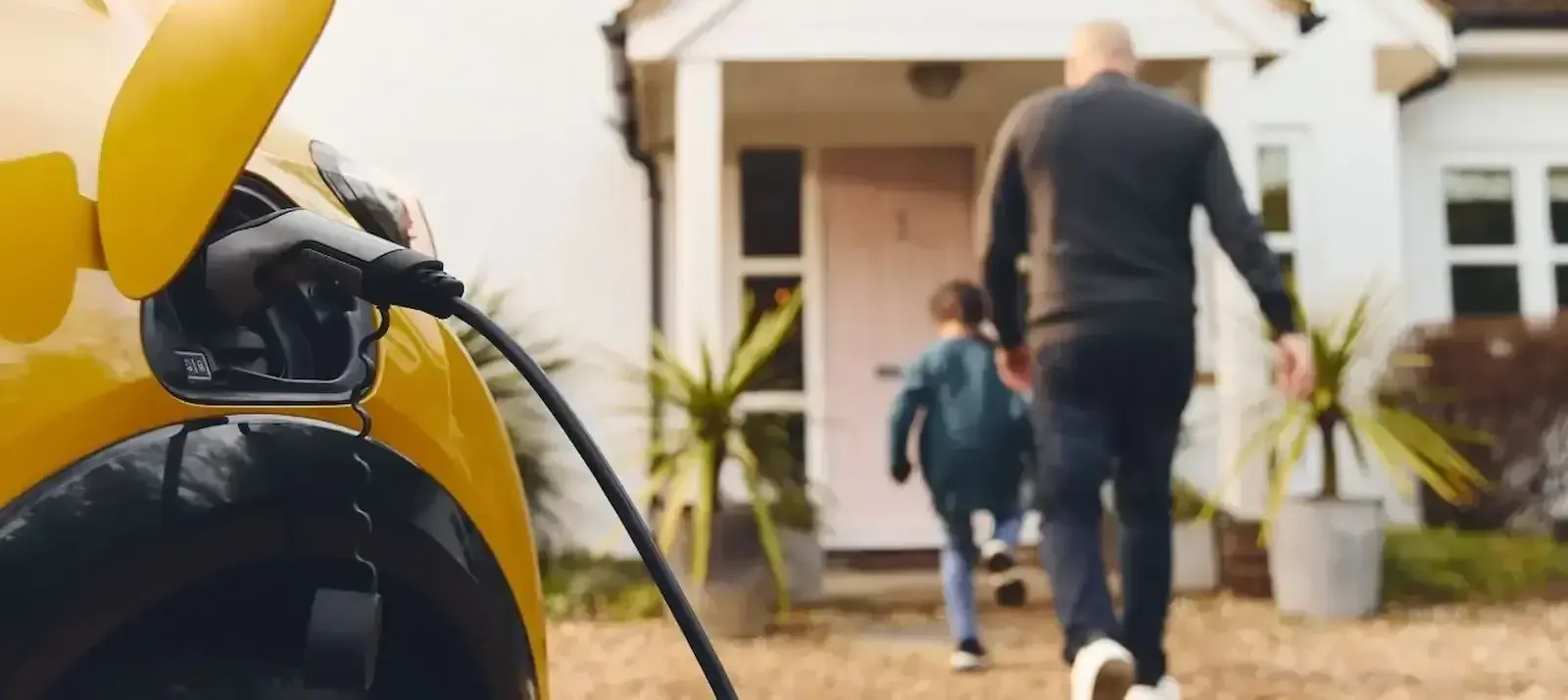A yellow electric car charging on a driveway as an adult and child walk up to the house