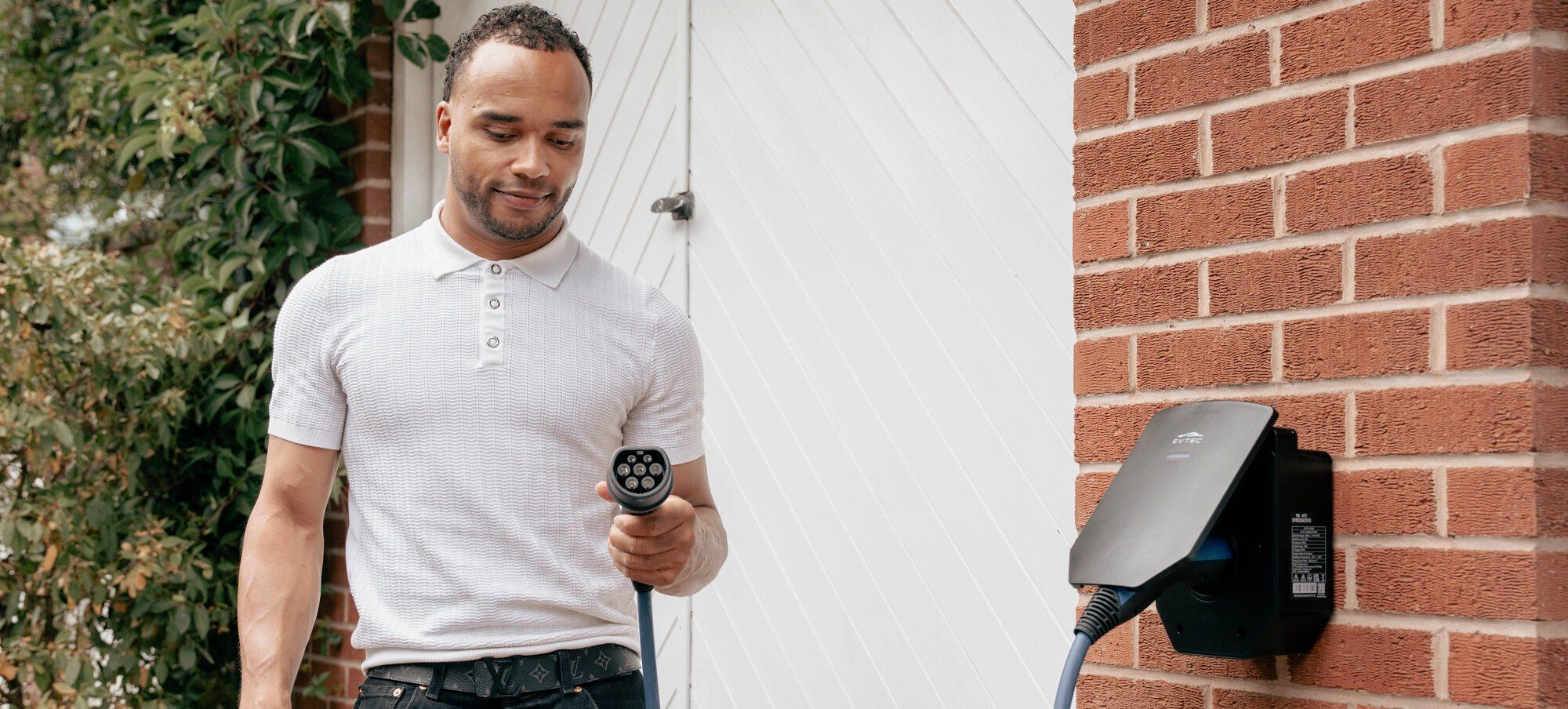 A man holding a charging connector beside a wall-mounted home charge point