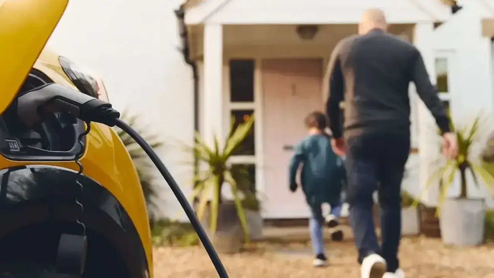 A yellow electric car charging on a driveway as an adult and child walk up to the house