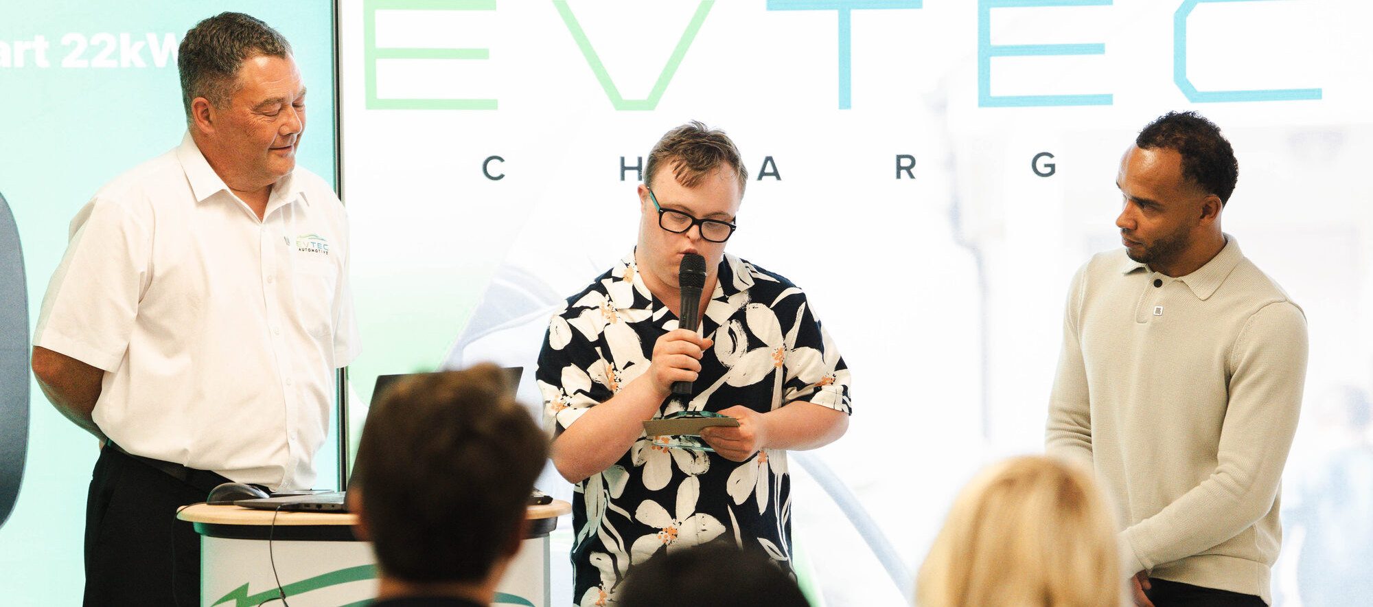 Three colleagues on stage at an Evtec Charge launch event, one speaking into a microphone